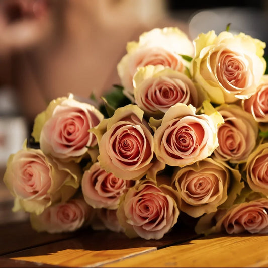 Buquê de rosas em tons pastel de rosa e amarelo em primeiro plano nítido, com mulher loira sorridente desfocada ao fundo em ambiente interno acolhedor. As rosas apresentam pétalas delicadas com gradações suaves de cor.