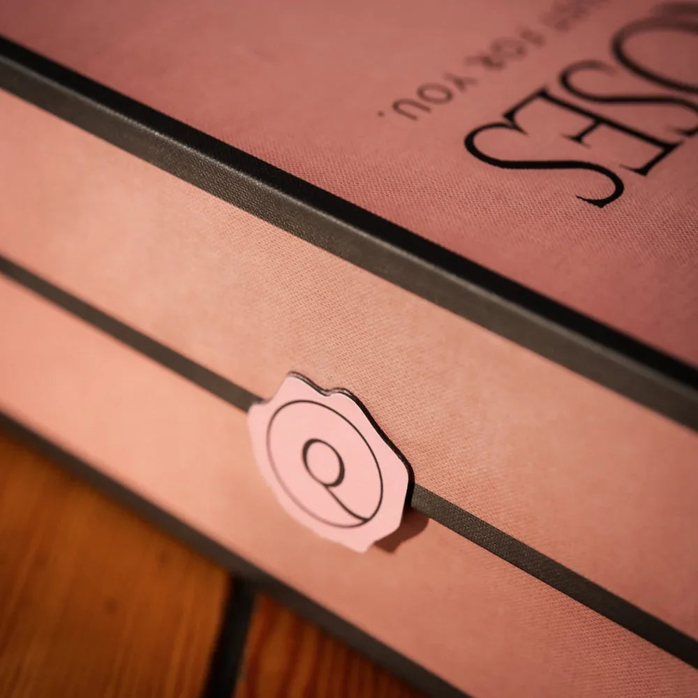 Close-up de caixa de presente elegante da Q_Roses em tom rosa com acabamento refinado, mostrando detalhe do lacre adesivo em formato de rosa e parte do logotipo da marca. A embalagem premium demonstra o cuidado artesanal na apresentação do produto.